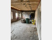 Ein leerer Innenraum in einem Gebäude im Rohbau, mit unverputzten Ziegelwänden, einem Betonboden und einer offenen Holzbalkendecke. Im Raum befinden sich Bauutensilien wie ein Eimer, Zementsäcke und gestapelte Ziegelsteine; zwei Personen stehen im hinteren Bereich und unterhalten sich.