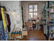 Ein kleines Geschäft mit Holzfußboden, dekoriert mit Regalen, die Keramik, Dekoartikel und Postkarten ausstellen. Links hängen bunte Kleidungsstücke auf einer Stange, während im Hintergrund ein großes Fenster den Raum erhellt.