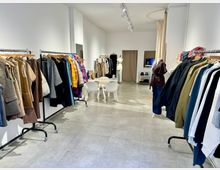 Ein moderner, heller Ladenraum mit zwei Reihen von Kleiderständern, die verschiedene Kleidungsstücke präsentieren, darunter Jacken, Mäntel und bunte Textilien. Im Hintergrund befinden sich ein Tisch mit Stühlen, ein Vorhang, und eine Schneiderpuppe sowie kleine Regale mit Accessoires.