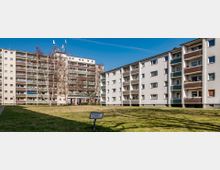 Mehrfamilienhäuser in einem Wohngebiet mit hellen Fassaden und Balkonen, umgeben von einer Rasenfläche. Im Vordergrund steht ein Hinweisschild, und vereinzelte Bäume säumen das Gelände.