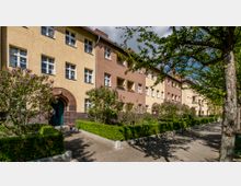 Mehrstöckiges Wohngebäude im Stil der 1920er-Jahre mit gelb-beigen und rötlichen Fassaden, dunkel abgesetztem Sockelbereich und Rundbogenportalen. Vor dem Gebäude verläuft ein Gehweg, gesäumt von Fliederbüschen, Hecken und Bäumen.