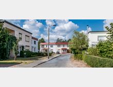 Wohnsiedlung mit modernen Einfamilienhäusern in heller Putzfassade und roten Dächern. Die Straße ist asphaltiert, von Grünflächen und Hecken gesäumt, und in der Mitte steht eine Straßenlaterne; im Hintergrund sind Bäume und ein blauer Himmel mit Wolken sichtbar.