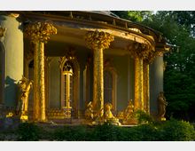Dekorativer Pavillon mit goldverzierten Säulen und filigranen Statuen in chinesischem Stil, umgeben von Bäumen und Pflanzen. Die Fassade zeigt hohe Fenster und verzierte Türen in einem elegant geschwungenen Aufbau.