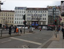 Straßenszene in einer Stadt mit mehrstöckigen Wohn- und Geschäftsgebäuden im Hintergrund, darunter eine moderne Glasfassade und klassische Fassaden mit Fenstern. Im Vordergrund sind eine Kreuzung mit Ampeln, Fußgänger, Fahrräder und ein orangefarbener Mülleimer erkennbar.
