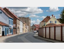 Straßenszene in einer Kleinstadt mit einer schmalen, asphaltierten Straße, gesäumt von niedrigen Wohnhäusern mit roten Ziegeldächern und teils bunten Fassaden auf der linken Seite. Rechts stehen Garagen mit weißen Wänden und Ziegelpfeilern, im Hintergrund einige Bäume und ein bewölkter Himmel.