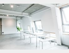 Minimalistisch gestalteter Innenraum mit weißen Wänden und schrägen Fenstern, die viel natürliches Licht hereinlassen. Im Vordergrund stehen weiße Tische und Bänke aus Metall, im Hintergrund ist eine grüne Zimmerpflanze zu sehen.