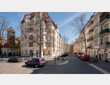 Eine gepflasterte Straße in einem Wohnviertel mit historischen Gebäuden im Stil der Gründerzeit sowie einem Kirchturm auf der linken Seite. Die Szenerie ist von Bäumen und parkenden Autos geprägt; im Vordergrund Fahrräder und öffentliche Sitzgelegenheiten.