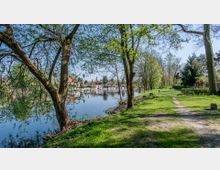Grüne Uferlandschaft an einem ruhigen Fluss, gesäumt von Bäumen und einem schmalen Fußweg. Im Hintergrund sind Häuser mit roten Dächern sowie eine Kirche mit einem Turm zu sehen.