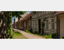 Schmale Wohnstraße mit Gehweg, gesäumt von niedrigen Häusern mit Satteldächern, teils als Fachwerkfassaden mit vielen Fenstern. Am Straßenrand stehen Gras und Bäume; die Gebäude liegen dicht aneinander und bilden eine geschlossene Häuserzeile.