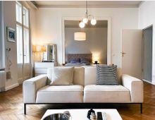 Helles Wohnzimmer mit einem cremefarbenen Sofa im Vordergrund, Holzboden, großen Fenstern auf der linken Seite und einer offenen Verbindung zu einem Schlafzimmer mit Doppelbett im Hintergrund. Eine Hängelampe und minimalistische Dekorationen ergänzen den modernen Stil.