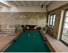 Ein rustikaler Raum mit sichtbaren Ziegelwänden und Holzbalkendecke, ausgestattet mit einem Billardtisch im Vordergrund. Im Hintergrund stehen eine Couch, ein Sessel, ein Holzregal und Wanddekorationen vor großen Fenstern mit grünen Fensterrahmen.