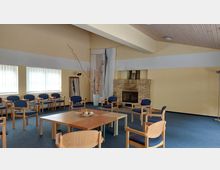 Ein heller Raum mit gelblichen Wänden, blauen Teppichboden und einer Sitzgruppe mit Holzstühlen und blauen Polstern, die um einen rechteckigen Holztisch arrangiert sind. An einer Wand steht ein gemauerter Kamin, daneben Dekorationselemente wie ein großer Spiegel und Zweige in einer Vase.