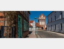 Enge gepflasterte Straße in einer Altstadt mit Fachwerkhäusern und historischen Gebäuden; im Hintergrund ein gotisches Backsteingebäude mit verzierten Giebeln unter blauem Himmel.