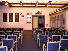 Ein kleiner Vortragssaal mit gepolsterten Stühlen in Reihen, einer beleuchteten Kassettendecke und einer Wand mit Fotos und Bildern. Rechts befinden sich dekorative Rundbögen im romanischen Stil, die an eine Galerie erinnern.