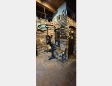 Rustikaler Innenraum mit freiliegenden Backsteinwänden, Ziegelboden und Holzdecke, mit einem gemauerten Rundbogen über einer Tür. Im Vordergrund steht eine große schwarze Handhebelpresse, daneben einfache Stühle; Regale und Wandflächen sind mit Flaschen und Vintage-Schildern dekoriert.