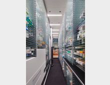 Kompakter Lagerraum in einer modernen Apotheke mit hohen Regalen, in denen zahlreiche Medikamentenschachteln aufgereiht sind. In der Mitte ein automatisierter Greifarm zum Sortieren und Entnehmen der Produkte.