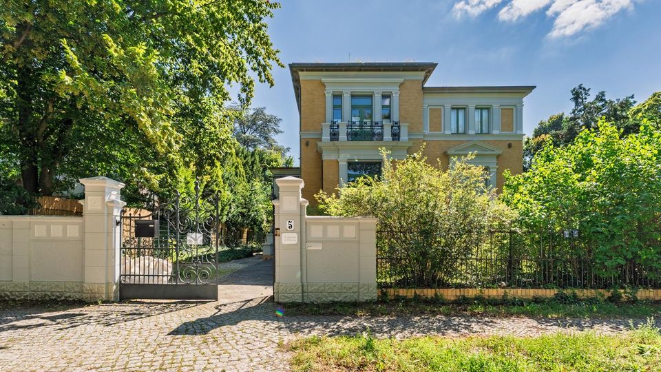 Zweistöckige Stadtvilla mit gelber Putzfassade, weißen Gesimsen und einem kleinen Balkon über dem Eingang, umgeben von Bäumen und Sträuchern. Vor dem Haus stehen eine niedrige Mauer und ein schmiedeeisernes Tor mit Zufahrt von einer gepflasterten Straße.