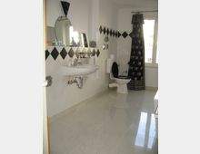 Helles Badezimmer mit glänzendem Fliesenboden, weiß-schwarzen Wandfliesen in Rautenmuster, einem Waschbecken, einem Wandspiegel, einer Toilette mit schwarzem Sitz und einer weiß-gemusterten Duschvorhang neben einem Fenster mit Tageslicht.