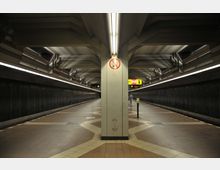 Unterirdischer U-Bahn-Bahnhof mit symmetrischem Design, gekennzeichnet durch eine zentrale Säule mit einem Rauchen-verboten-Schild, flankiert von zwei Bahnsteigen und dunklen Wänden. Überkopf sind beleuchtete Deckenelemente und Informationsschilder sichtbar.