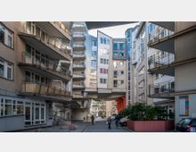 Innenhof mit modernen Wohngebäuden, deren Fassaden teilweise aus Beton, Ziegel und Metall bestehen, mit zahlreichen Balkonen und großen Fenstern. Im Vordergrund Gehweg mit Fußgängern, parkenden Autos und Pflanzenbeeten.