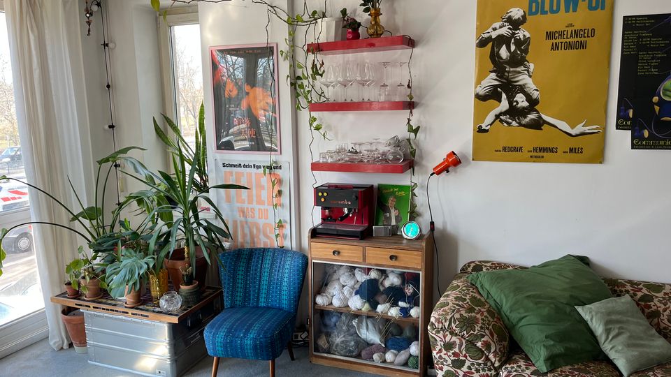 Cozy living room corner beside tall windows with sheer curtains, featuring several potted plants on a low trunk, a blue upholstered chair, and a floral-patterned sofa with green cushions. A small cabinet with shelves of glassware and a coffee machine sits against a white wall decorated with trailing vines, string lights, and large vintage film posters.