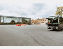 Großer asphaltierter Platz mit einem modernen Gebäude mit Glasfassade auf der linken Seite und einem zweistöckigen Backsteingebäude mit mehreren Fenstern im Hintergrund. Ein schwarzer Bus steht rechts im Bild, und orangefarbene Absperrungen sind auf dem Platz verteilt.