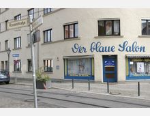 Straßenszene mit einem Gebäudeeckladen mit der Aufschrift „Der blaue Salon“, einem Friseursalon mit blauer Tür und dekorierten Fenstern. Im Vordergrund sind Straßenbahnschienen, ein Straßenschild an einer Laterne sowie ein geparkter Wagen zu sehen.