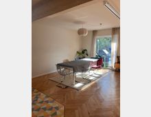 Esszimmer mit einem rechteckigen Tisch, umgeben von verschiedenen Stühlen, auf einem Teppich mit Holzboden in Fischgrätenmuster. Große Fenster mit transparenten Vorhängen lassen Tageslicht herein, neben Pflanzen im Raum. Im Vordergrund farbig gemusterte Fliesen.