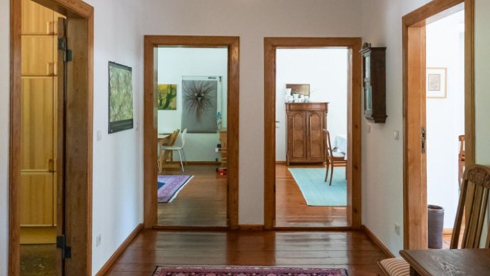 Heller Flur in einer Wohnung mit Holzdielenboden, orientalischem Läuferteppich und zentraler Pendelleuchte. Mehrere Türöffnungen mit Holzrahmen führen in angrenzende Räume; rechts steht ein Holztisch mit Stuhl, links eine Glastür.