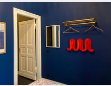 Blick in ein Zimmer mit dunkelblauer Wand, weiß gerahmter offener Tür und kleinem Wandspiegel. Daneben hängen eine goldfarbene Garderobenstange mit Kleiderbügeln und eine rote, geschwungene Wandgarderobe.