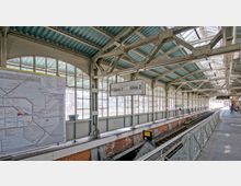 Bahnhofshalle mit verglasten Wänden und einem grünen Wellblechdach. Im Vordergrund ein Liniennetzplan, ein Schild mit den Gleisangaben "Gleis 2" und "Gleis 3", sowie Bahngleise darunter; im Hintergrund Sitzbänke und eine digitale Anzeigetafel.