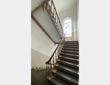 Historisches Treppenhaus mit dunklen Holzstufen, einem verzierten Holzgeländer und einer großen, bogenförmigen Fensteröffnung, die Tageslicht in den Raum lässt. Die Wände sind in hellen Beige- und Weißtönen gehalten.