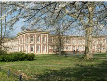 Ein historisches Gebäude mit rot-weißer Fassade und großen Rundbogenfenstern, umgeben von einem Park mit Bäumen und grüner Wiese. Im Vordergrund sind blühende Zweige und ein Fußweg zu sehen.
