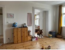 Wohnzimmer eines Hauses mit Holzdielenboden und weißen Wänden. Im Vordergrund steht eine holzfarbene Kommode mit einem Globus darauf, daneben ein Spielzeugpferd und gestapelte Spielzeugkisten. Rechts ist ein großes Fenster mit gelben Vorhängen, im Hintergrund führt eine offene Tür in ein weiteres Zimmer mit einem Tisch und Stühlen.