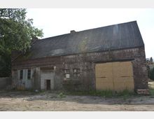 Backsteinbau mit Wellblechdach, teils beschädigt, mit zugemauerten Fenstern und einer großen Holztür. Die Umgebung wirkt verwildert, mit Erde und Graswuchs.