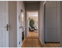 Flur in einer Wohnung mit Holzboden, weißen Wänden und einer Türöffnung, die in ein Wohnzimmer führt. Im Hintergrund sind ein großer Zimmerpflanze, eine helle Couch und ein Teppich sichtbar, rechts ein moderner Einbauschrank.