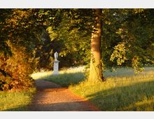 Ein bewaldeter Parkweg mit von Sonnenlicht beleuchteten Bäumen und Wiesen; im Hintergrund steht eine weiße Steinskulptur auf einem Sockel.