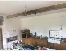 Küche mit rustikalen Elementen, darunter eine freiliegende Holzbalkendecke, Holzschränke und eine weiße Spüle mit Fliesenrückwand. Mehrere Küchenutensilien und Gegenstände sind auf den Arbeitsflächen verteilt.