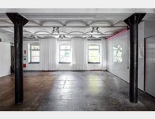 Industrieähnlicher Innenraum mit hohen Decken und zwei freistehenden schwarzen, metallenen Säulen. Drei Fenster mit weißen Vorhängen lassen Tageslicht herein, der Boden besteht aus abgenutztem Beton, und rechts ist eine glänzende, weiße Wand erkennbar.