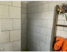 Eckdusche mit weißen Wandfliesen in rechteckigem Format, daneben eine dekorative Holzleiter mit einem orangefarbenen Handtuch und getrockneten Blumen.