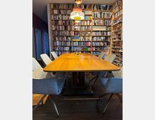 Ein Raum mit einer großen Regalwand voller Bücher, einem hölzernen rechteckigen Tisch in der Mitte und mehreren grauen Polsterstühlen. Über dem Tisch hängen farbige Pendelleuchten, der Raum hat einen Holzboden.