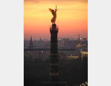 Die Siegessäule in Berlin bei Sonnenuntergang, mit der goldenen Figur der Viktoria auf ihrer Spitze. Im Hintergrund sind städtische Gebäude, Kräne und ein farbenfroher Himmel in Orange- und Rottönen zu sehen.