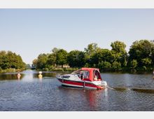 Ein kleines Motorboot mit rotem Sonnenschutz fährt einen ruhigen Fluss entlang, umgeben von Bäumen und grüner Vegetation. Im Hintergrund ist eine blaue Brücke zu sehen, und der Fluss ist markiert mit rot-weißen Tonnen.