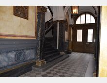 Historisches Treppenhaus mit Marmorsäulen, aufwendig gestalteten Wandverzierungen und einem Holztüren-Eingang mit Glasfenstern im Hintergrund. Der Boden ist mit einem geometrischen Muster gefliest, und eine Treppe mit dunklen Stufen führt nach oben.