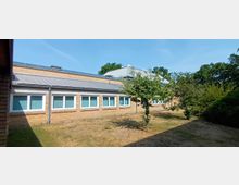 Flacher Backsteinbau mit mehreren rechteckigen Fenstern, teilweise von Bäumen und Sträuchern umgeben, auf einer Rasenfläche. Das Gebäude hat ein hellgraues Metalldach mit Solarpanels und befindet sich in einer grünen Umgebung.