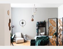 Modern eingerichtetes Wohnzimmer mit hellem Holzfußboden, grauer Wand und minimalistischen Möbeln. Zu den Details gehören ein beiger Sessel, ein kleiner Beitisch, eine dekorative Wanduhr, hängende Lampen, ein Paravent mit goldfarbenen Blattmustern und eine Regalkonstruktion mit grüner Decke und Accessoires.