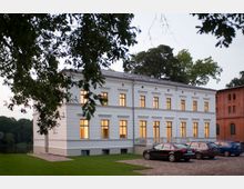 Zweistöckiges weißes Gebäude mit symmetrischen Fenstern, deren Innenlichter leuchten, umgeben von Bäumen und einer Wiese. Im Vordergrund befinden sich gepflasterte Parkplätze mit mehreren geparkten Autos; rechts steht ein rotes Ziegelgebäude.