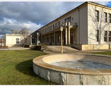 Zweistöckiges, hellbeiges Gebäude mit Säulen am Eingangsbereich, großen rechteckigen Fenstern und einem umliegenden Grünbereich. Im Vordergrund befindet sich ein leerer, runder Brunnen aus Stein.