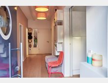 Flur eines modernen Wohnraums mit heller Holzbodenoptik, einer offenen Tür, an der ein rundes Fenster eingelassen ist, und mehreren roten Deckenlampen. An der rechten Wand befinden sich weiße Schränke und Regale, daneben ein roter Stuhl, während an der linken Wand Hüte an Haken hängen.