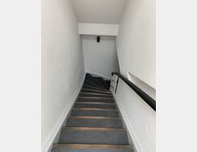 Geschwungene Treppe in einem engen Treppenhaus mit weißen Wänden, grauem Bodenbelag und Holzleisten an den Treppenstufen; an der rechten Wand befindet sich ein schwarzer Handlauf, und an der Stirnwand hängt eine wandmontierte Leuchte.